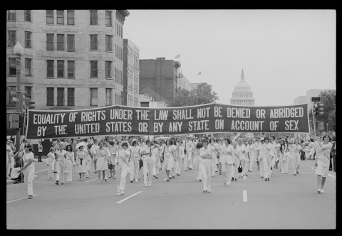 Women wearing all white marching in the street and carrying a banner that says "Equality of rights under the law shall not be denied or abridged by the United States or by any state on account of sex"