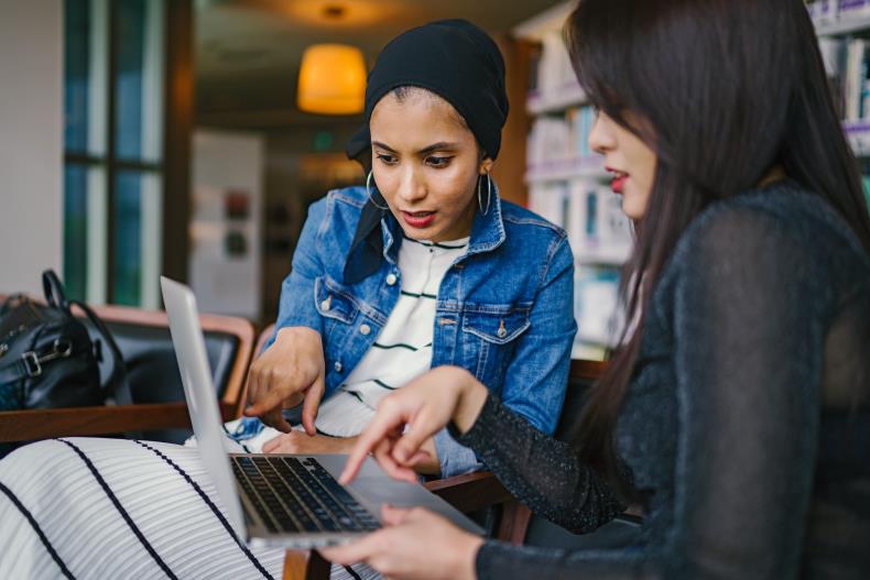 two women sitting at a desk, looking at a laptop