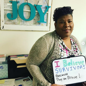 Woman holding up a dry erase board that says "I believe survivors because they are brave!"