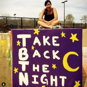 Woman sitting on top of a large concrete block on which "Take Back the Night," stars, and a moon are painted