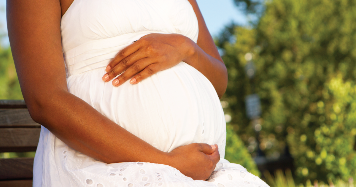 Close-up of a pregnant Black woman's belly. She is wearing a white dress and cradling her stomach.
