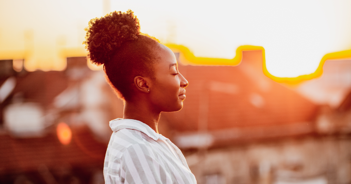 Black woman facing a sunset with her eyes closed, meditating