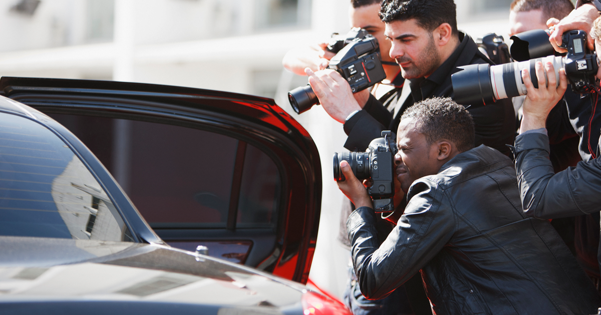 A limousine door is open, surrounded by multiple paparazzi snapping pictures of the celebrity inside