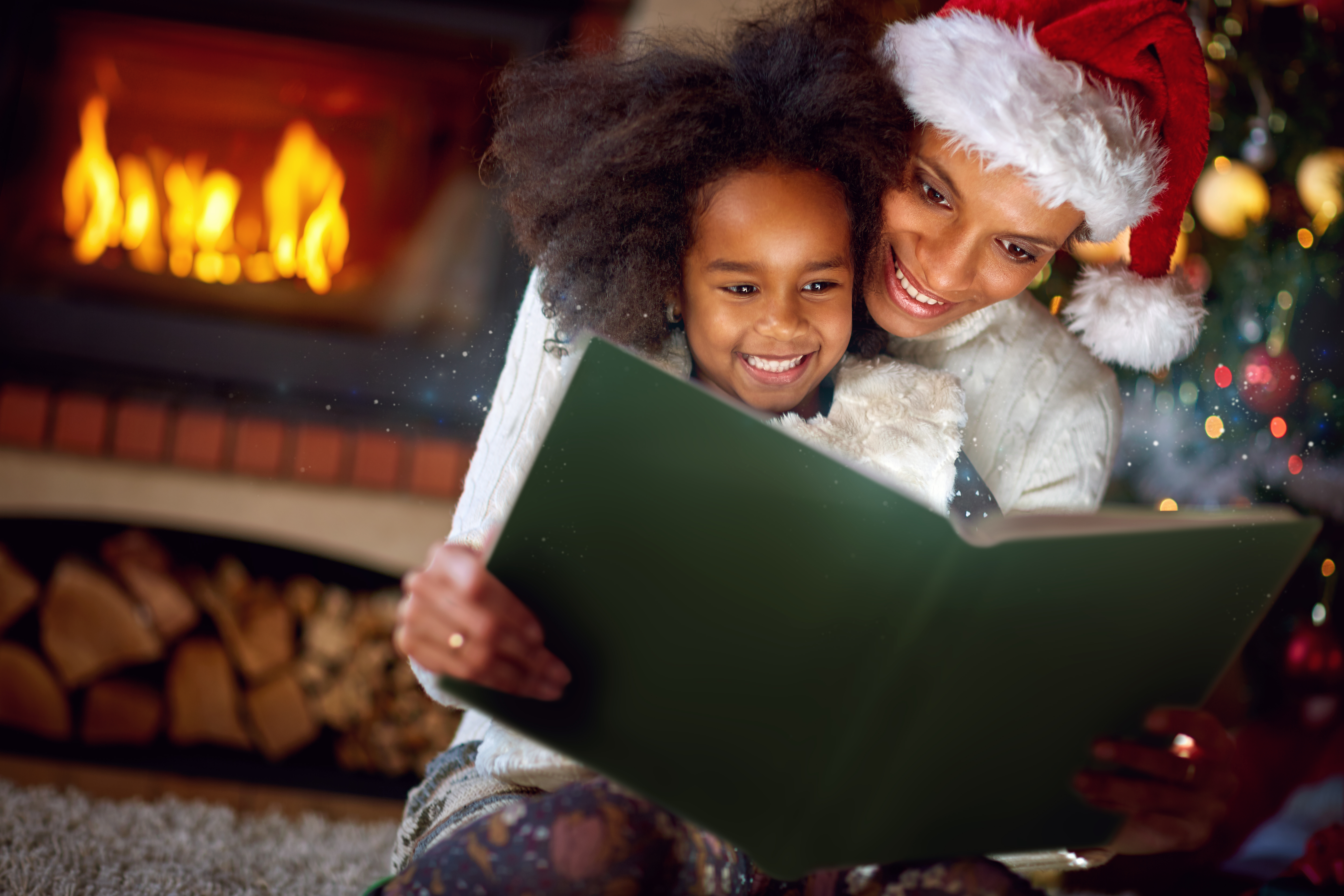 a happy child reads a book in the arms of a loved one (seemingly her mother). The adult is wearing a santa hat and they are sitting in front of the fire. Their faces are lighting up from a light beaming from the insight of the book.