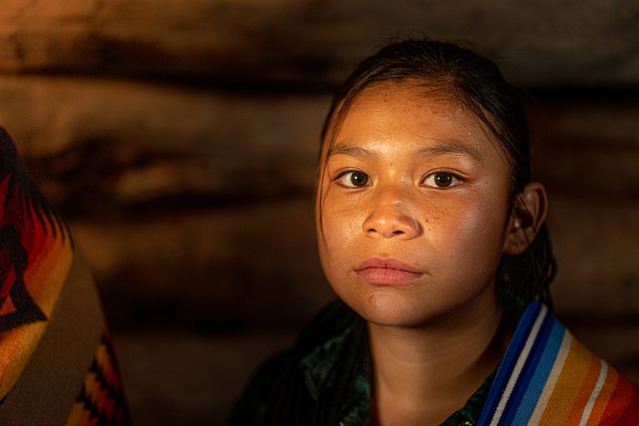 Image is a portrait of a native girl looking seriously directly at the camera