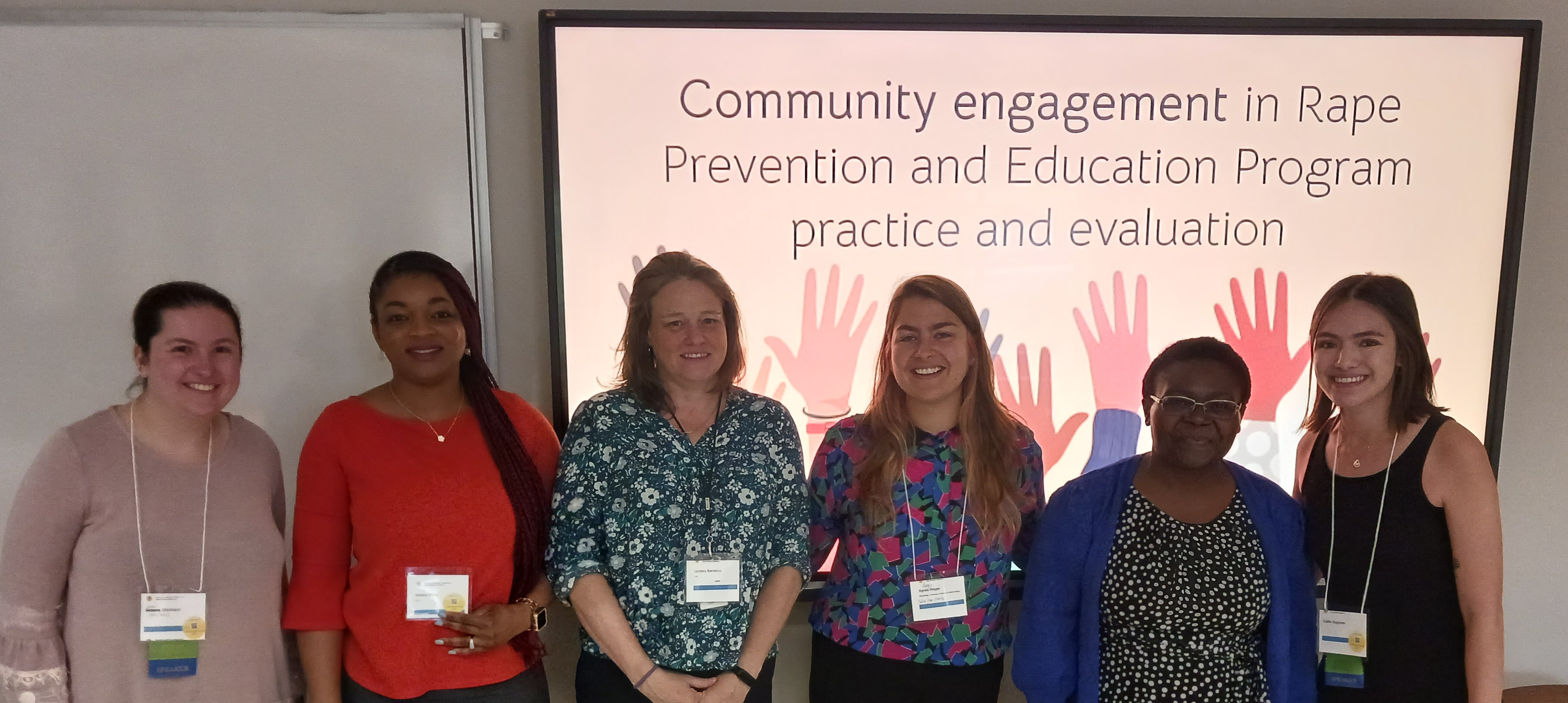 This is a photo of the in-person presenters at the Community engagement in Rape Prevention and Education panel, at the Society for Community Research and Action. The photo is of a group of people, standing side by side and smiling. They are wearing business-casual clothes and conference name badges. Behind them is a presentation slide that reads: "Community engagement in Rape Prevention and Education."  