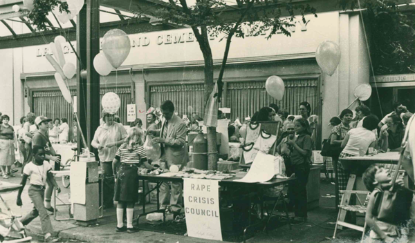 People gathered around a table at a community event with a sign that reads "Rape Crisis Council"