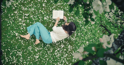 Young woman lying down on the grass outside reading, surrounded by flowers petals