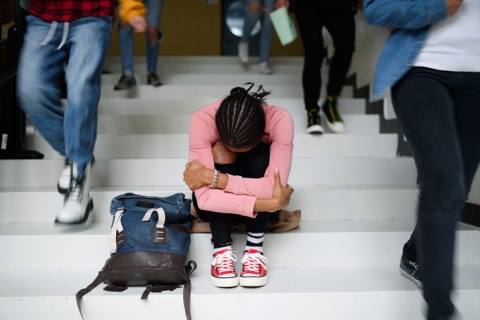 image shows the steps of a school, with a student hunched over sadly with their head between their knees and their backpack infront of them as other students pass by them down the stairs. 