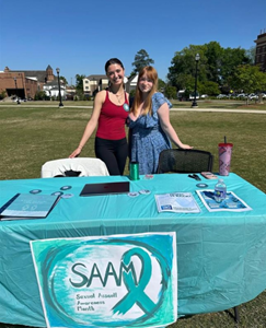 Two women standing together behind SAAM table